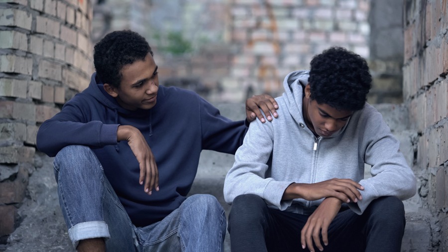 Young man comforting teenager, both sitting on a staircase in an alleyway with brick walls