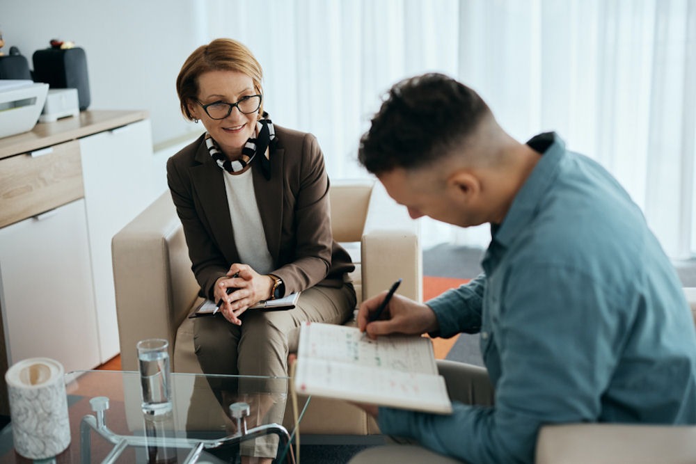 Man taking notes on his anxiety and addiction during a session with therapist wearing glassestherapist taking notes in a notebook